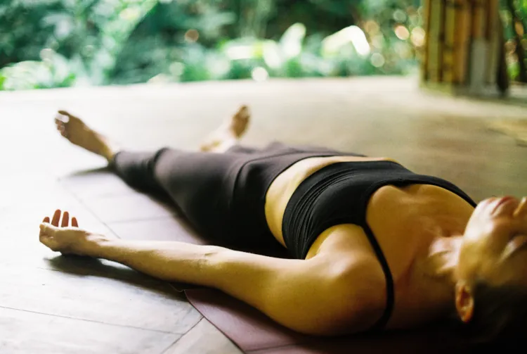 Person in black workout attire lies in a peaceful, relaxed position on a yoga mat, surrounded by lush greenery, conveying calm and tranquility.