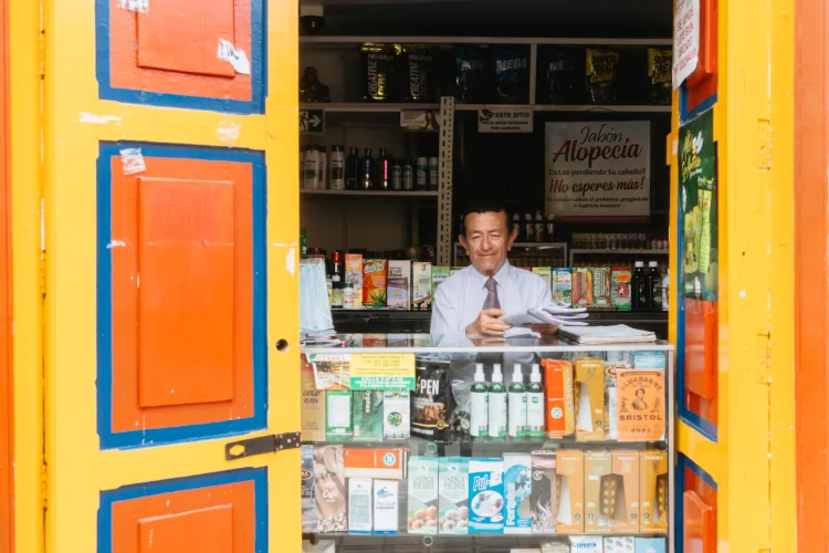 Man in a shop stands behind a counter filled with various products. The open red and yellow door frames his smiling face, creating a welcoming atmosphere.