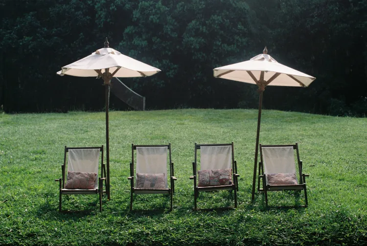 Two white umbrellas shade four wooden lounge chairs with patterned cushions on a green lawn, bordered by lush trees, evoking a serene outdoor setting.