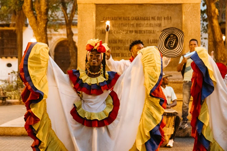 A smiling dancer in vibrant traditional attire with red, yellow, and blue ruffles performs energetically. Background shows a lively street scene.