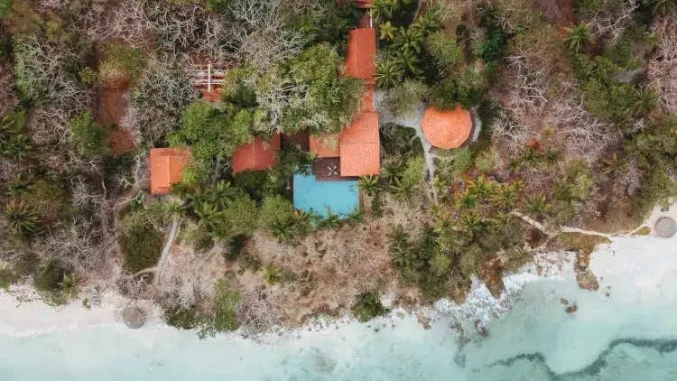 Drone overhead of secluded tropical villa with red-tiled roofs, turquoise pool, palm trees, dense green jungle, and private beachfront on Caribbean coastline