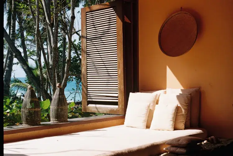Woman in black swimsuit and rust headscarf drinking from fresh coconut while lounging on wooden beach chair with ocean backdrop