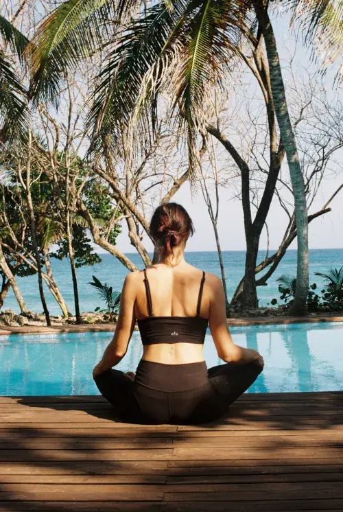 Woman in black Alo yoga outfit meditating in lotus pose on wooden deck overlooking infinity pool and palm-fringed Caribbean sea