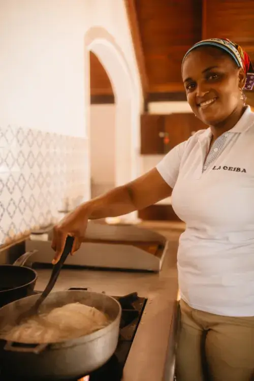 Smiling woman chef in La Ceiba polo shirt stirring pot in open-air kitchen with arched stone walls, wooden beams, and tiled counters