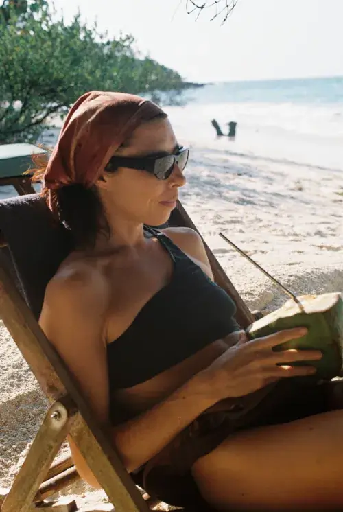 Woman in black swimsuit and rust headscarf drinking from fresh coconut while lounging on wooden beach chair with ocean backdrop