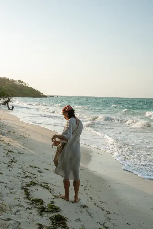 Woman in light linen dress and headscarf strolling barefoot on sandy beach with turquoise ocean waves and palm trees at golden hour