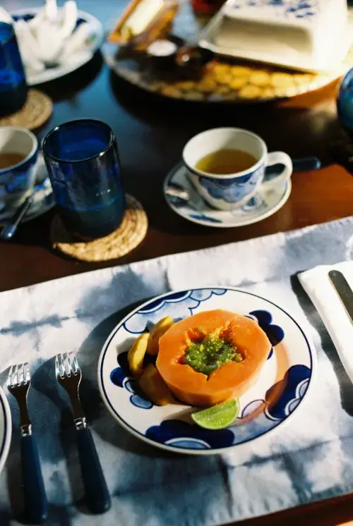 Blue and white patterned plate with halved papaya topped with green salsa, lime wedge, and tropical fruit slices on woven coaster