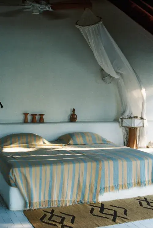 Rustic bedroom with adobe-style plastered walls, mosquito net canopy over bed with blue and beige striped bedding, shelf with terracotta vases and wooden figurine, ceiling fan, and natural sunlight