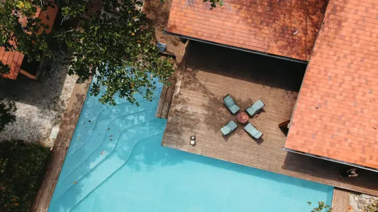 Aerial shot of turquoise swimming pool with wooden deck featuring circular seating arrangement, red-tiled roof, surrounding greenery, and small table setup in Mediterranean-style villa courtyard