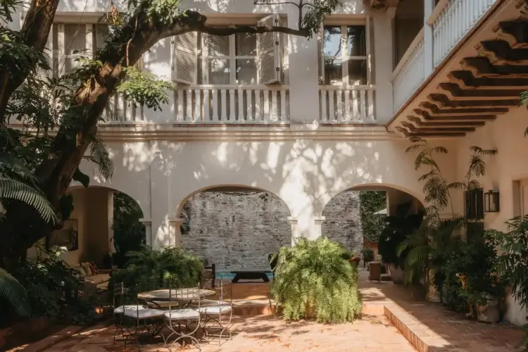 Serene courtyard with arches, lush greenery, and dappled sunlight. Features a round table, ferns, and a view of a bright, inviting swimming pool.