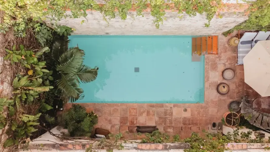Overhead view of a serene courtyard pool surrounded by lush greenery and terracotta tiles. A sun umbrella and cozy seating area add a relaxed vibe.