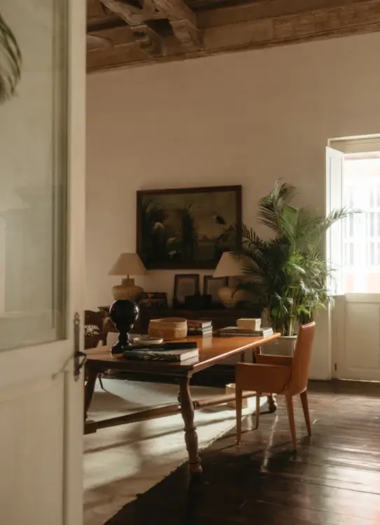 Cozy study with elegant wooden desk, tan chair, and green plants by an open window. Warm light, vintage decor, and framed art create a serene ambiance.