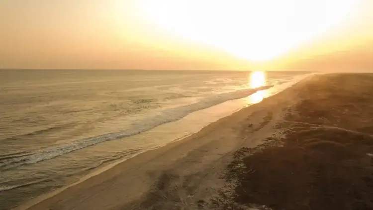 Wide-angle shot of a pristine, sandy beach and rolling ocean waves under a hazy golden sunset.