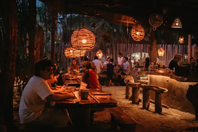 A cozy, evening restaurant scene with sand floors and heavy wooden tables. Warm, circular woven lanterns hang from the ceiling, illuminating groups of people dining and talking.