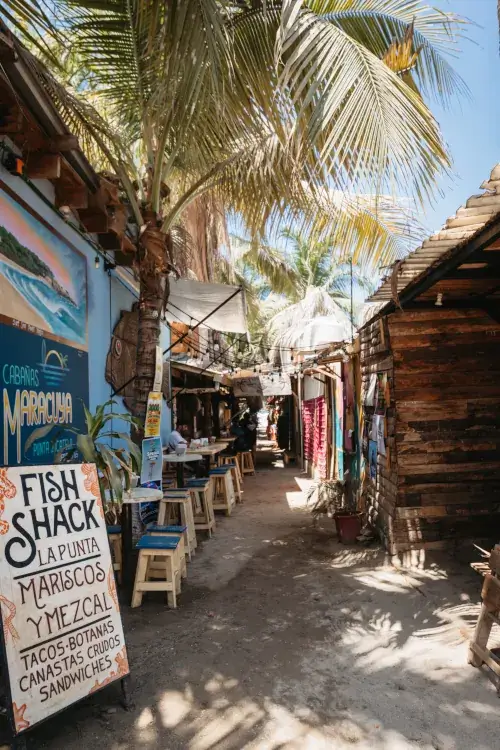 A narrow sandy corridor between rustic buildings and palm trees. A hand-painted sign for 