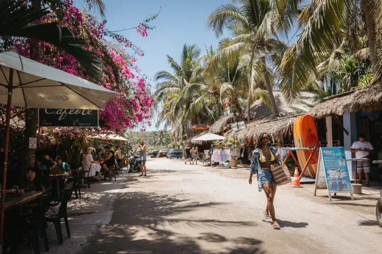 A wide, sandy street in a beach town lined with palm trees and thatched-roof shops. A woman in denim shorts and a yellow bikini top walks toward the camera.