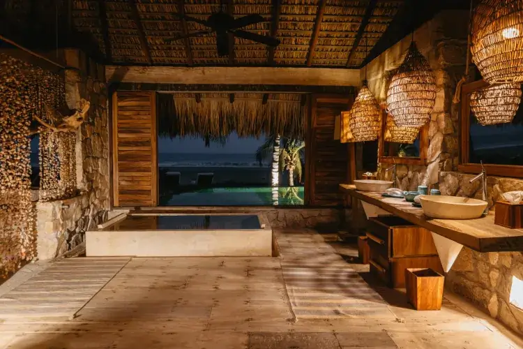 A dimly lit, high-end rustic bathroom with stone walls, wooden floors, and woven pendant lights. A large open window frames a view of a lit swimming pool and the ocean at dusk.