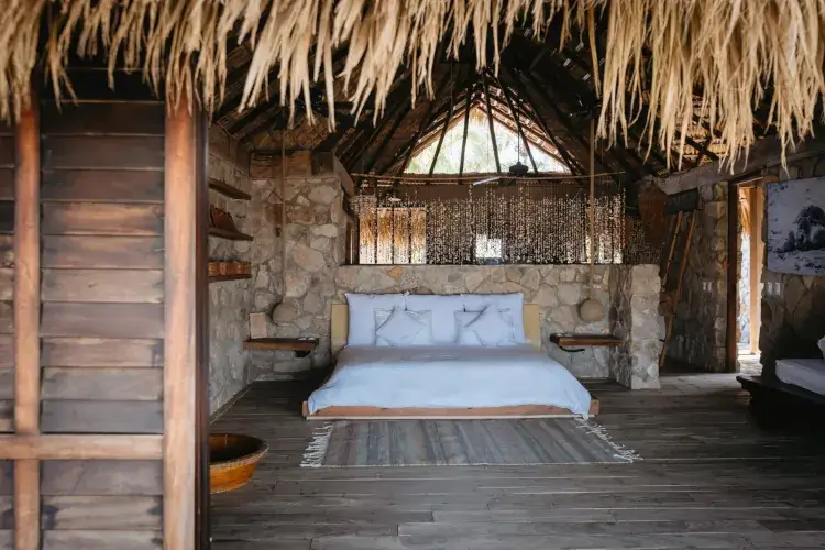 A rustic, open-air luxury bedroom in Puerto Escondido, Mexico, featuring a stone wall headboard, a thatched palapa roof, and a low wooden platform bed with crisp white linens. Two unique pendant lights hang over floating wooden nightstands, and a beaded curtain separates the sleeping area from the background.