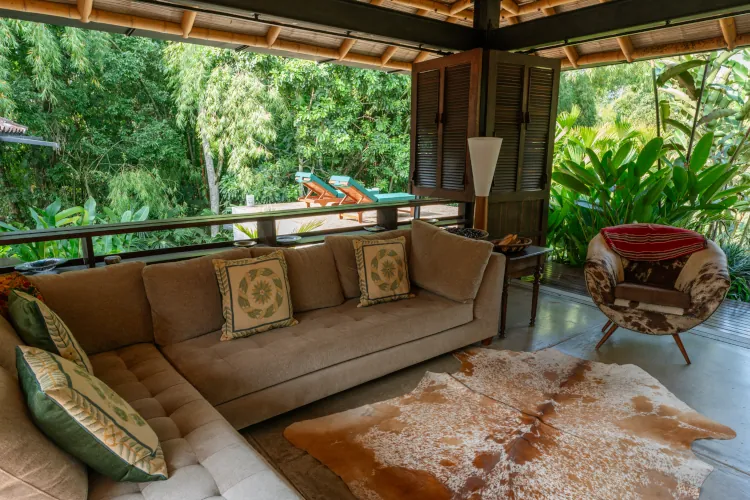 Cozy living room with a beige sofa, patterned cushions, and a cowhide rug. Wooden shutters open to a lush, green jungle view, creating a serene, tropical feel.