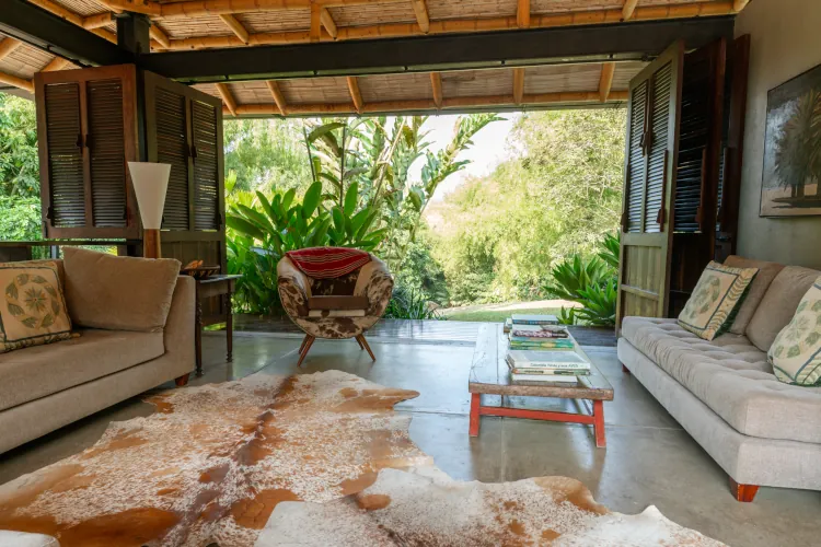 Open-air living room with beige sofas and a patterned armchair. A cowhide rug lies on concrete flooring. Lush greenery is visible outside through wide-open wooden shutters, creating a serene and inviting ambiance.