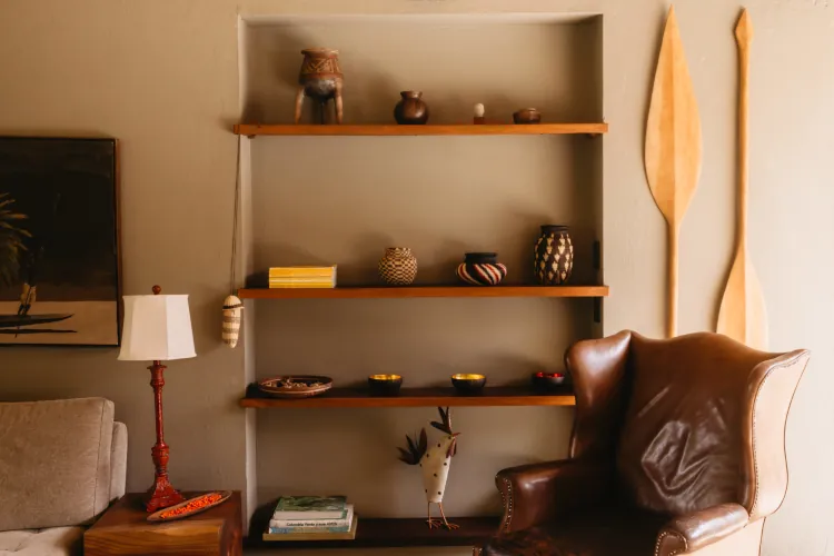 A cozy living room with wooden shelves adorned with pottery and books. A leather chair sits beside a lamp on a table, framed by decorative paddles.