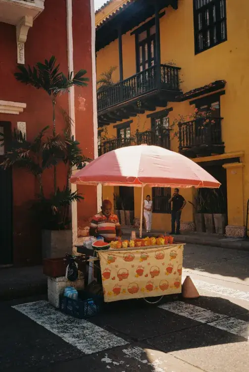 Street vendor with a colorful cart, under a pink umbrella, selling fruit on a sunny street corner. Two people stand by vibrant yellow and red buildings.