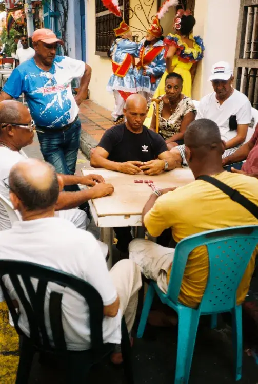 A group of men playing dominoes at a table on a vibrant street, with colorful costumes and smiling onlookers in the background. The scene feels lively and communal.