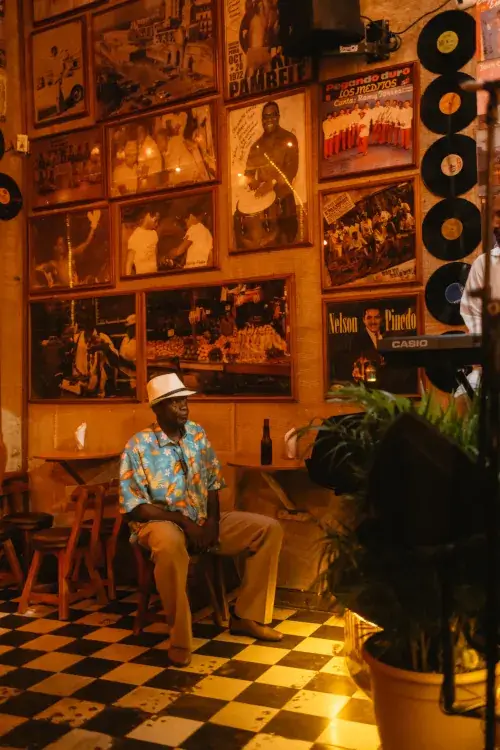 A man in a colorful shirt and hat sits relaxed on a chair in a warm, vintage-style bar with checkered floor. Walls are adorned with framed photos and records.