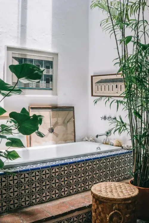 A serene bathroom with a tiled bathtub surrounded by lush green plants and framed art on white walls. The room exudes a calm, natural vibe.