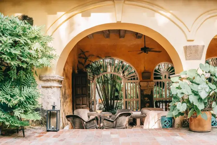 Sunny Spanish-style patio with arches, wicker chairs, and lush plants. A rustic fan hangs above, evoking a cozy, relaxed atmosphere.