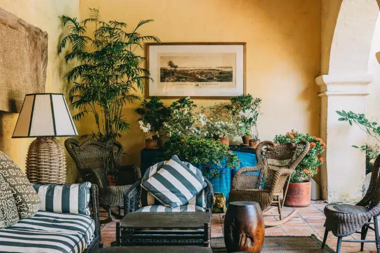 Cozy patio setting with wicker chairs, striped cushions, and a woven lamp. Lush potted plants and a framed painting create a warm, inviting atmosphere.