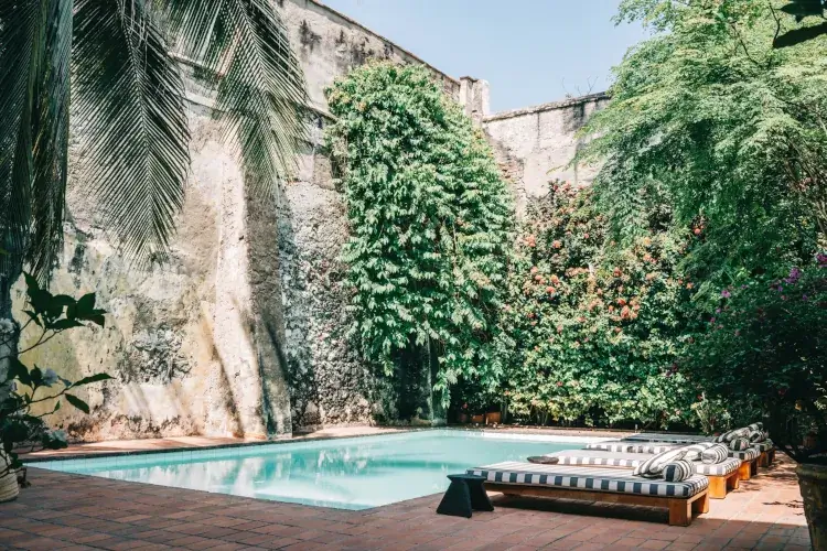 Secluded courtyard with a small pool and striped loungers. Surrounded by lush green plants and flowers, set against rustic, sunlit walls. Calm ambiance.