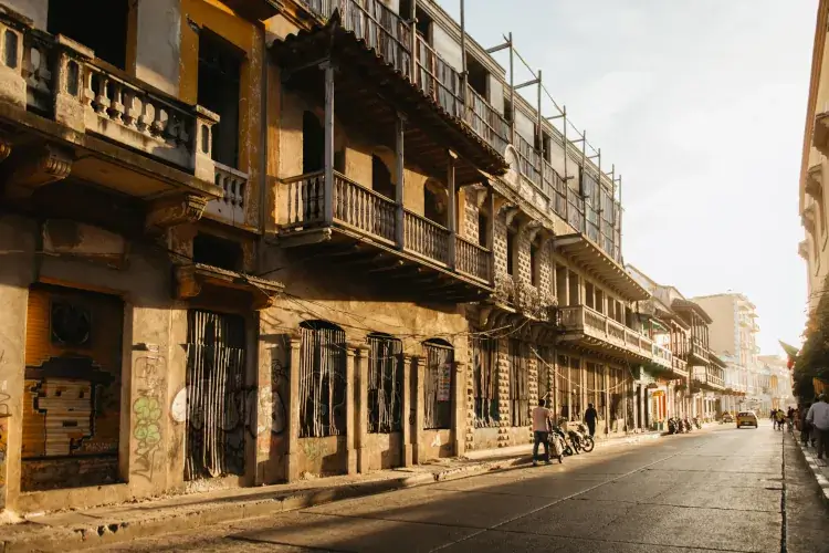 Street scene in warm sunlight with old, weathered buildings featuring balconies and graffiti. Few people walk along the quiet road, evoking a peaceful atmosphere.