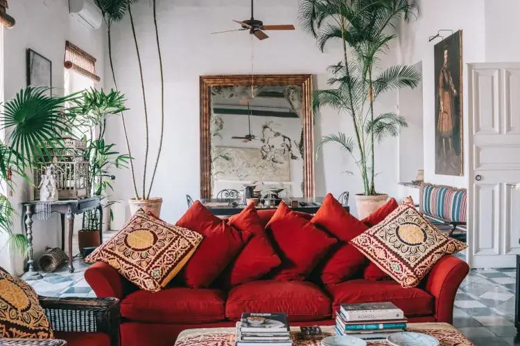 A vibrant living room with a red couch adorned with patterned pillows, lush green plants, a large mirror, and artistic decor, creating an inviting, eclectic atmosphere.
