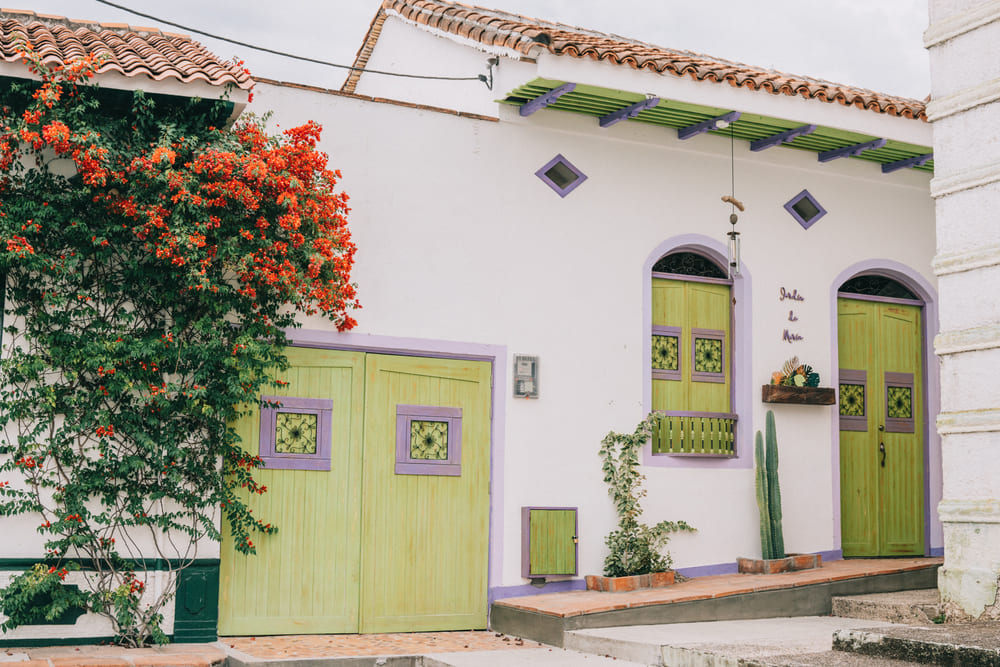 A charming white colonial house with bright lime-green doors and purple trim, surrounded by flowering plants and tropical greenery