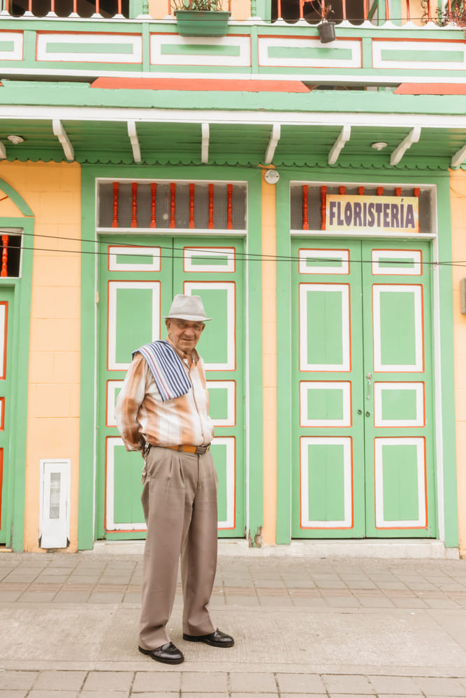 An older local man standing in front of a brightly painted green-and-yellow colonial building with a “Floristería” sign