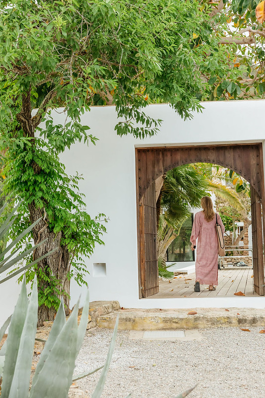 Woman walking through a wooden doorway into a white villa surrounded by greenery in Ibiza