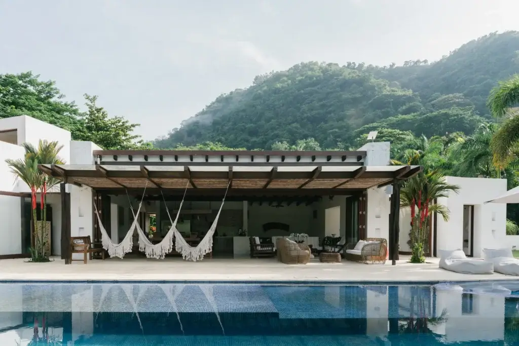 A luxury infinity pool in front of a covered terrace featuring three white fringed hammocks and outdoor seating.