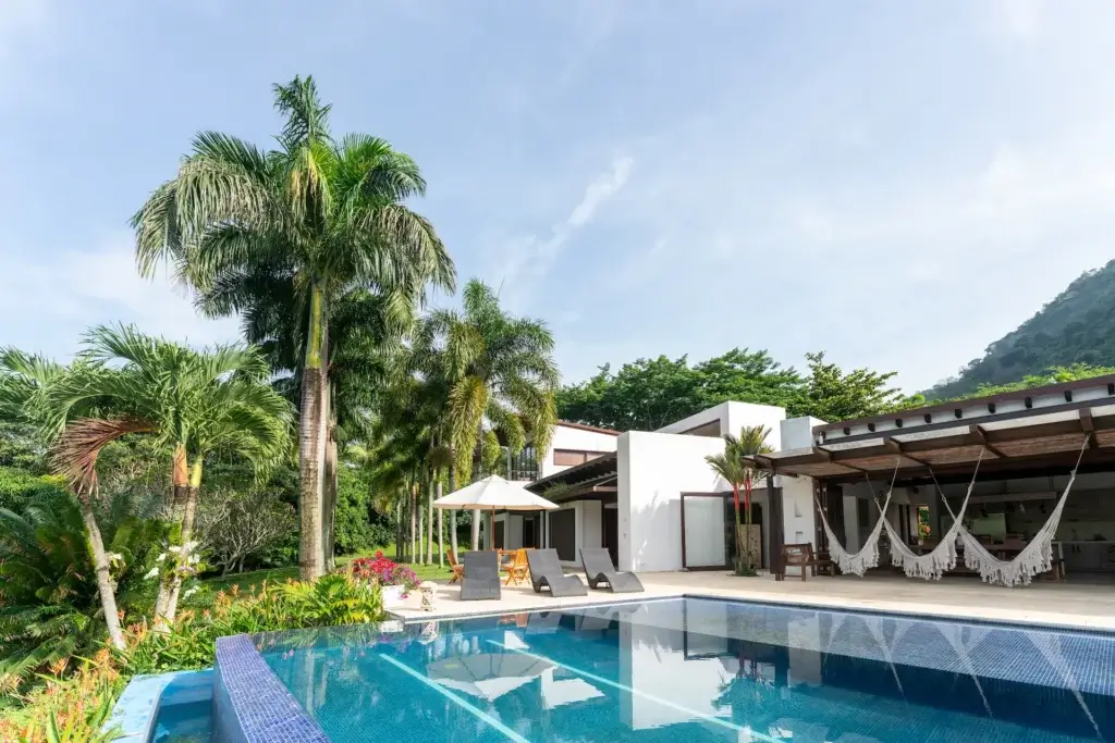 Three dark grey lounge chairs and a patio table set beside a swimming pool, shaded by tall palm trees.