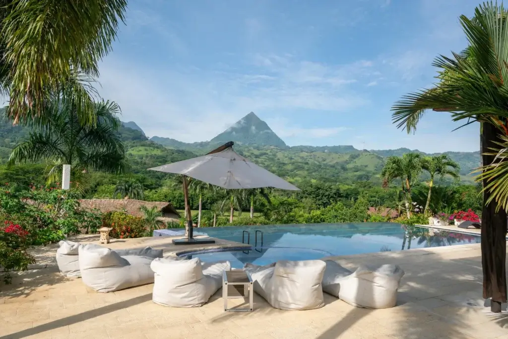: A large grey sun umbrella and white beanbag chairs by an infinity pool overlooking a valley and a pyramid-shaped mountain.