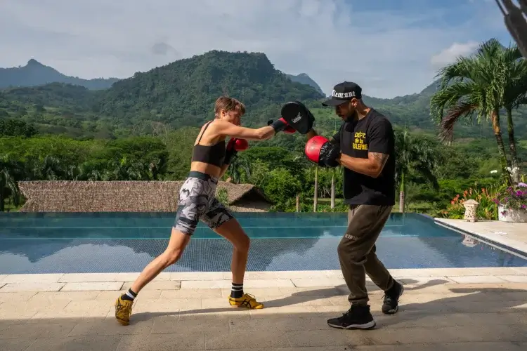 A person in athletic wear practices boxing techniques outdoors, with a scenic mountain backdrop and a swimming pool nearby.