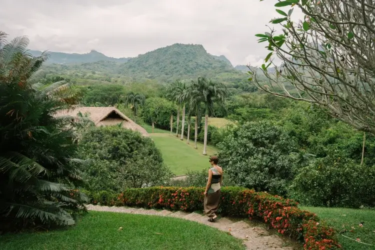 A person walks along a garden path, surrounded by lush greenery and mountains under a cloudy sky. A thatched roof structure is visible nearby.