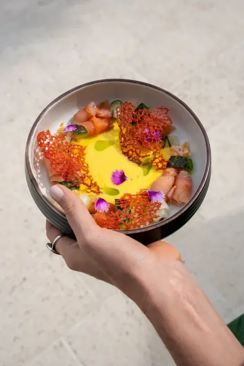A hand holds a bowl of vibrant, artistic sushi adorned with colorful toppings, including flowers and crispy elements, against a stone background.