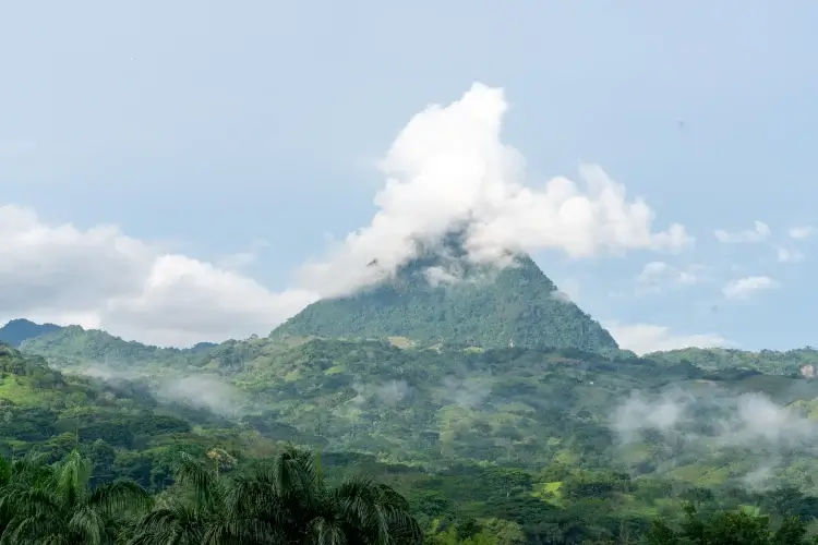 A daylight view of a steep, green-covered mountain peak with a large white cloud partially obscuring its summit. The foreground shows a dense, tropical forest valley under a pale blue sky.