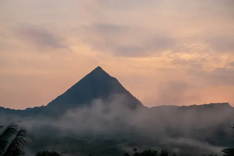 A wide shot of a distinct, pyramid-shaped mountain peak silhouetted against a soft orange and pink sunset sky. Thick white mist and low-lying clouds drift across the base of the mountain and the surrounding dark green valley.