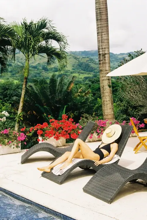 A person in a black swimsuit and a large straw hat relaxes on a grey wicker lounge chair by a pool. Bright pink bougainvillea flowers, palm trees, and rolling green hills create a tropical backdrop under a bright, overcast sky.