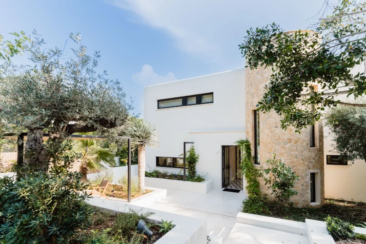 Modern house with a minimalist design, featuring white walls and vertical stone accent. Surrounded by lush greenery and trees under a clear blue sky.