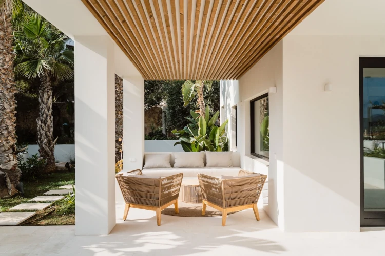 Covered patio with wooden slat ceiling, featuring two wicker chairs and a cushioned bench around a circular table. Surrounded by lush greenery and palm trees.