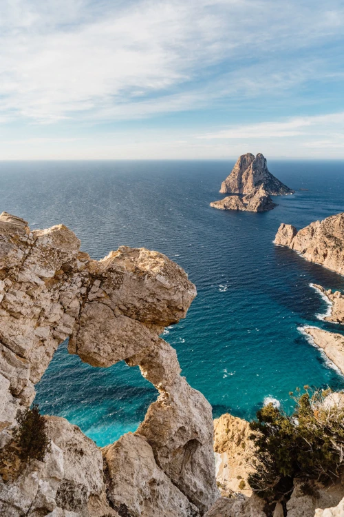 A rocky cliff with a natural arch frames a distant island surrounded by the blue ocean under a partly cloudy sky, creating a serene coastal scene.