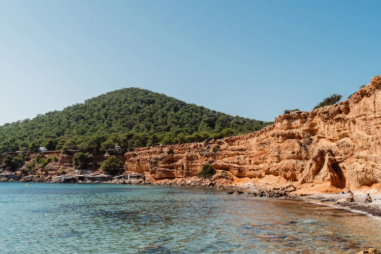 Sunny coastal scene with clear blue water, red rocky cliffs, and a green forested hill in the background. Calm and serene atmosphere.